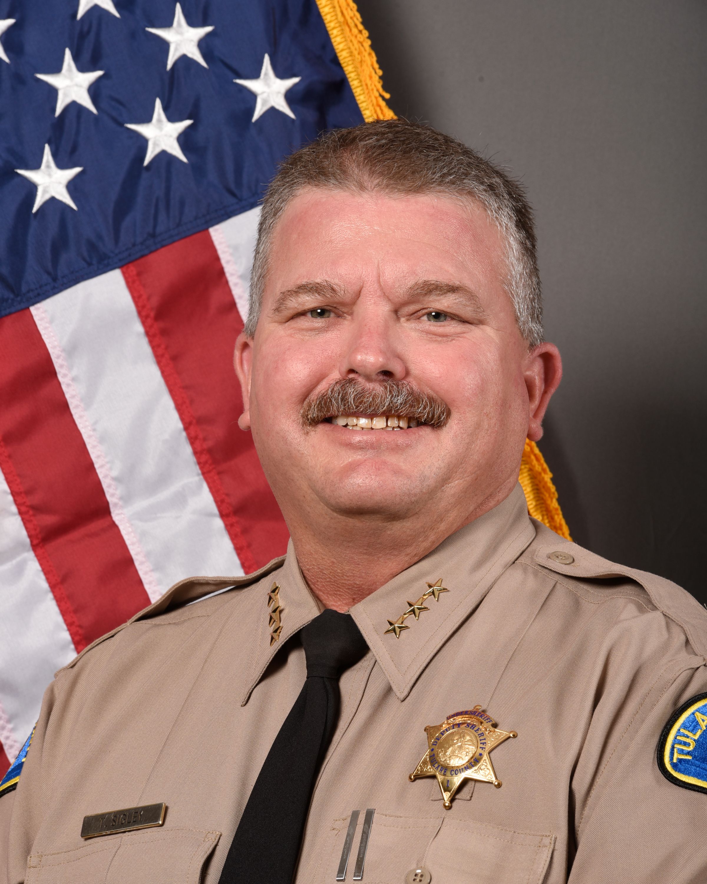 A portrait of Undersheriff Tom Sigley in uniform standing in front of an American Flag. 